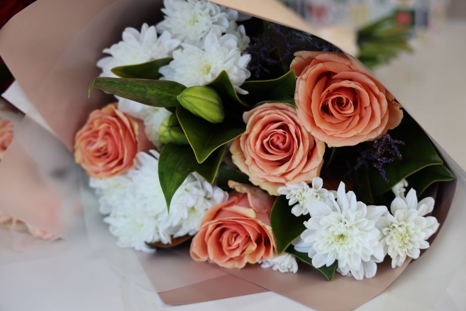 A floral arrangement featuring three peach-colored roses with gently ruffled petals, complemented by clusters of white chrysanthemum flowers with numerous layered petals. The bouquet also includes gre