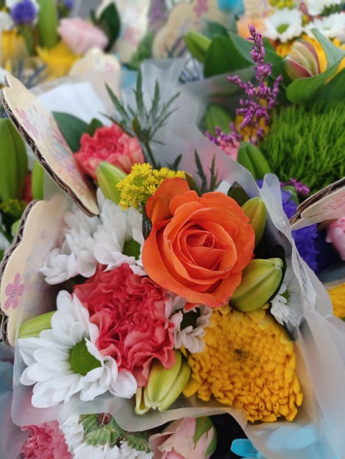 A vibrant floral arrangement featuring an orange rose as the focal point, surrounded by pink carnations, white daisies with green centers, yellow chrysanthemums, and purple lavender sprigs. The bouque