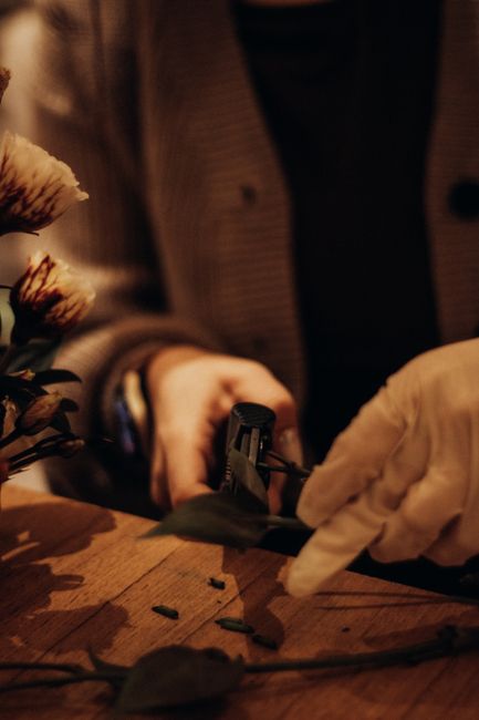 A close-up image of a florist at work, focusing on their hands as they carefully arrange a floral bouquet. The florist is dressed in a brown checked shirt with rolled-up sleeves and gloves, working ov