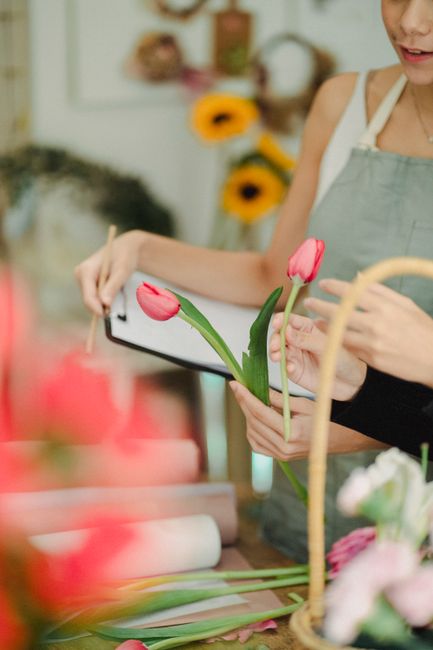 A florist from Cranford florists, wearing a light grey apron, is arranging a floral gift with two pink tulips held in her left hand and a smartphone in her right hand, possibly for reference. The work