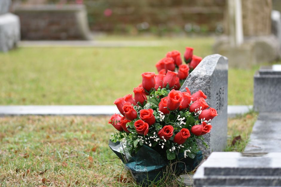A bouquet of fresh, vibrant red roses with tightly closed buds and lush green foliage, arranged in a rounded style with white baby's breath accents. The bouquet is wrapped in dark green floral paper a