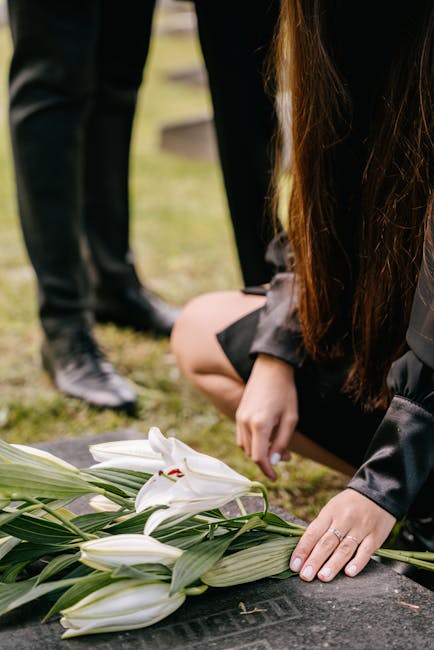 A person wearing black clothing is kneeling on the ground, gently placing a bouquet of white lilies with green stems and leaves onto a memorial stone. The lilies have large, open petals with a hint of
