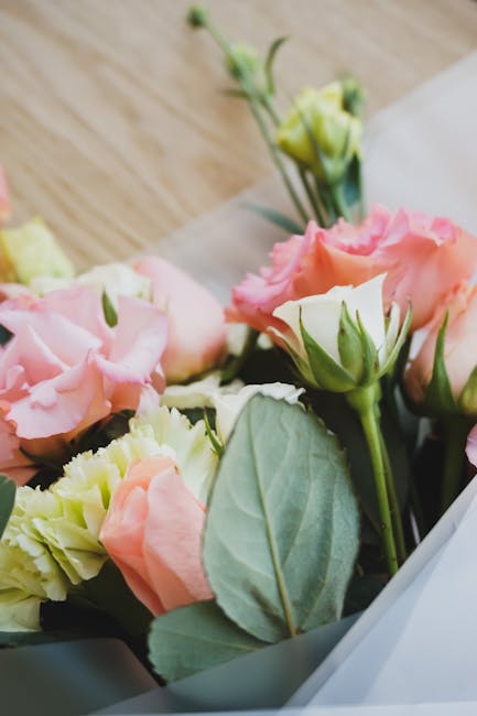 A close-up of a floral arrangement featuring pale pink and cream lisianthus flowers with ruffled petals, complemented by green foliage and greyish-green leaves. The flowers are freshly cut, with smoot