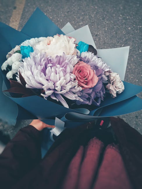 A hand holding a beautifully arranged floral bouquet wrapped in blue and white paper, featuring a mix of white chrysanthemums, soft pink roses, purple carnations, and bright blue accents. The flowers 