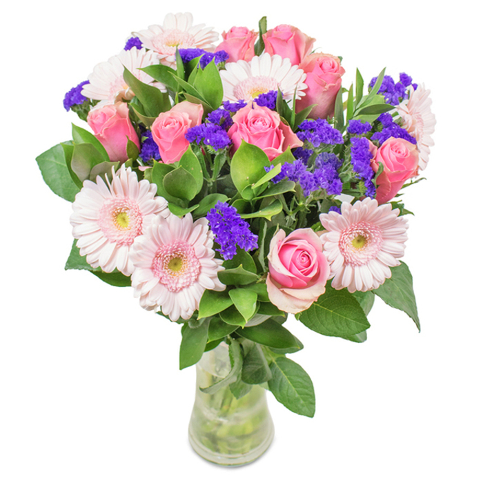 An eye-catching floral arrangement, hand-designed in Cranford and displayed against a luminous white background, features an enchanting balance of approximately 7-9 pink, spiral-centred roses-many still tightly furled beside others just opening to reveal intricate whorls. These are beautifully contrasted by around 5-7 pale pink gerbera daisies, their generous, radiating petals rippling out from near-white centres with a texture as plush as the morning light on Cranford's High Street. Throughout, clusters of vivid purple statice or limonium provide a striking pop of colour and a slightly papery texture, enhancing the tactile richness and visual depth of the bouquet. Deep green foliage fills the base of the clear glass vase, lending a lush, grounded feel and suggesting freshness akin to the wild greenery of local parks. The arrangement sits with a sense of poised abundance and contemporary grace, perfect as a fresh delivery for a Cranford birthday, Mother's Day, or as a warm welcome in an office overlooking the River Crane. Each element is chosen as much for sentiment as for beauty-an invitation to celebrate, nurture, or simply to brighten one's day with flowers crafted by local experts.